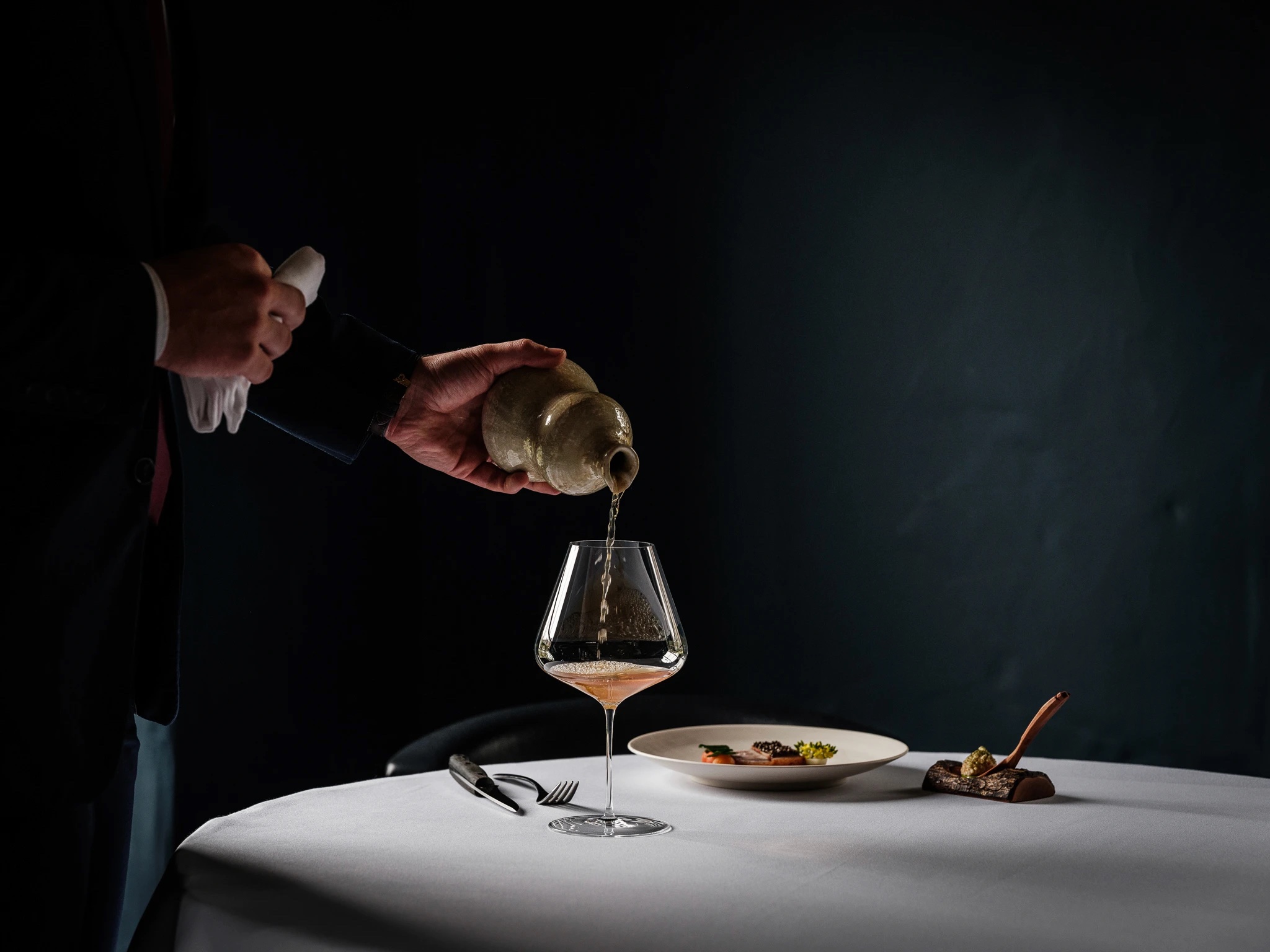 A sommelier pouring from a ceramic vessel into a glass at a fine-dining table