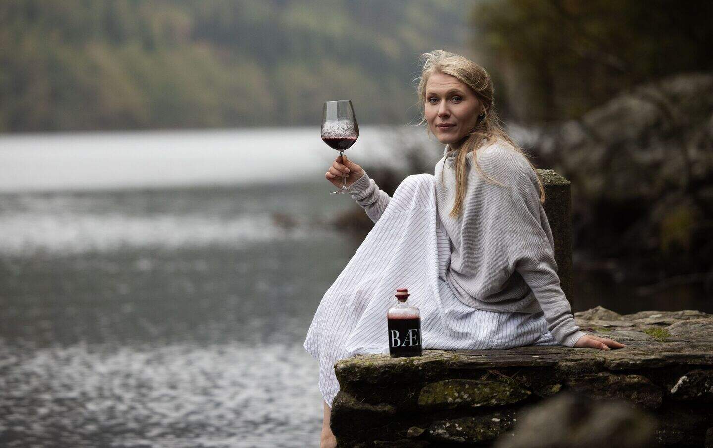 Majken Bech-Bailey by a lake with a glass and bottle of BÆ, her non-alcoholic wine alternative